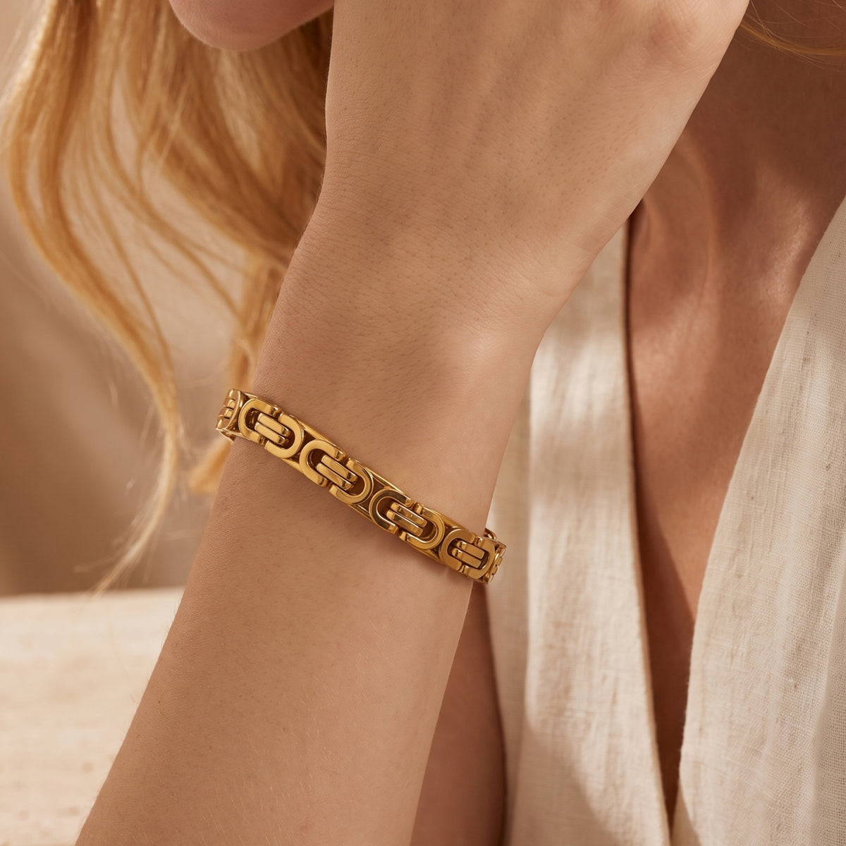 Gold-toned Byzantine link bracelet worn on a wrist beside cream-colored fabric.