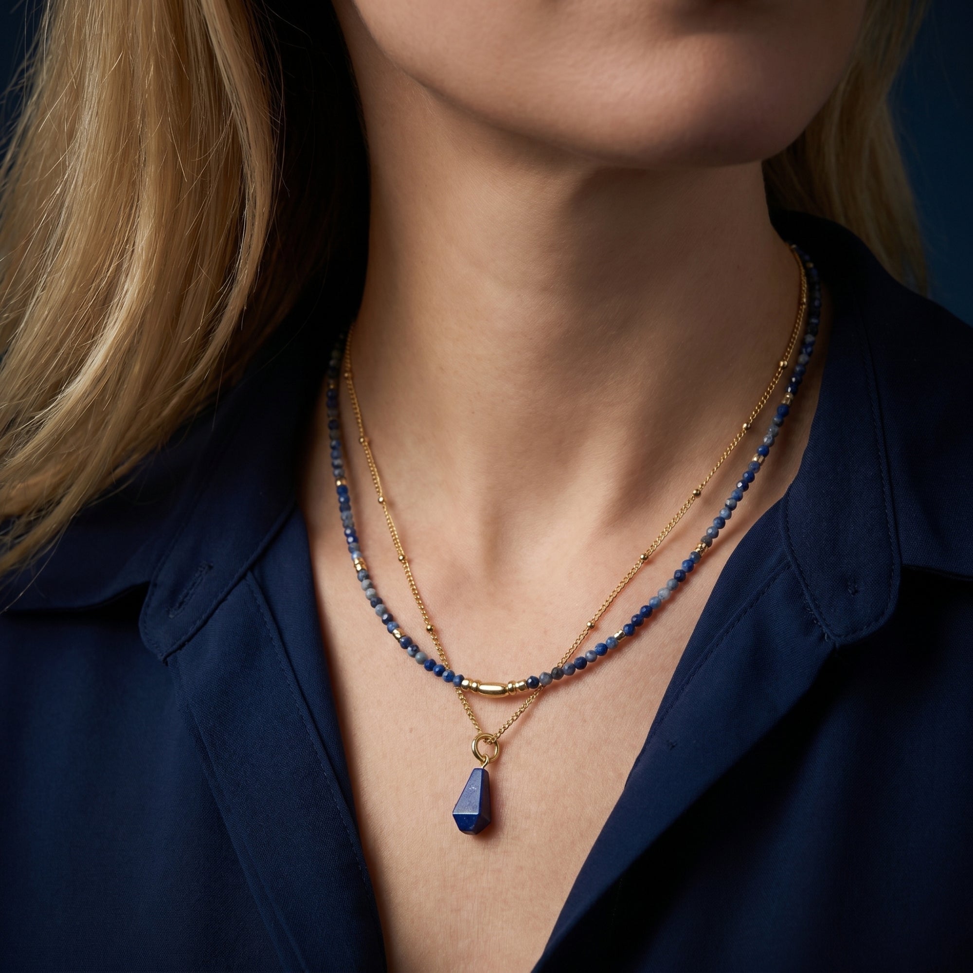 Gold double-layered necklace with natural lapis beads and pendant worn by a model in a navy blue shirt.