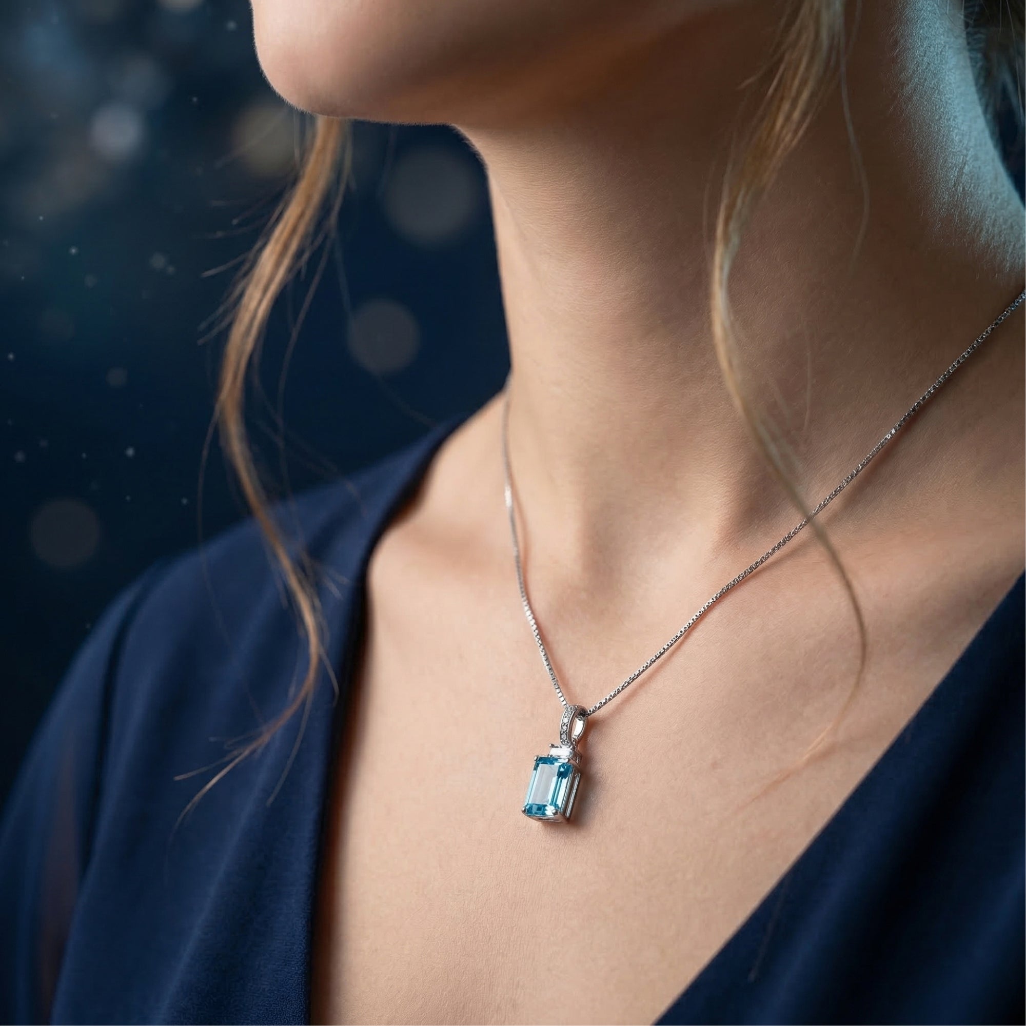 Side view of a model wearing an emerald cut blue topaz pendant on a silver chain with a dark navy dress.