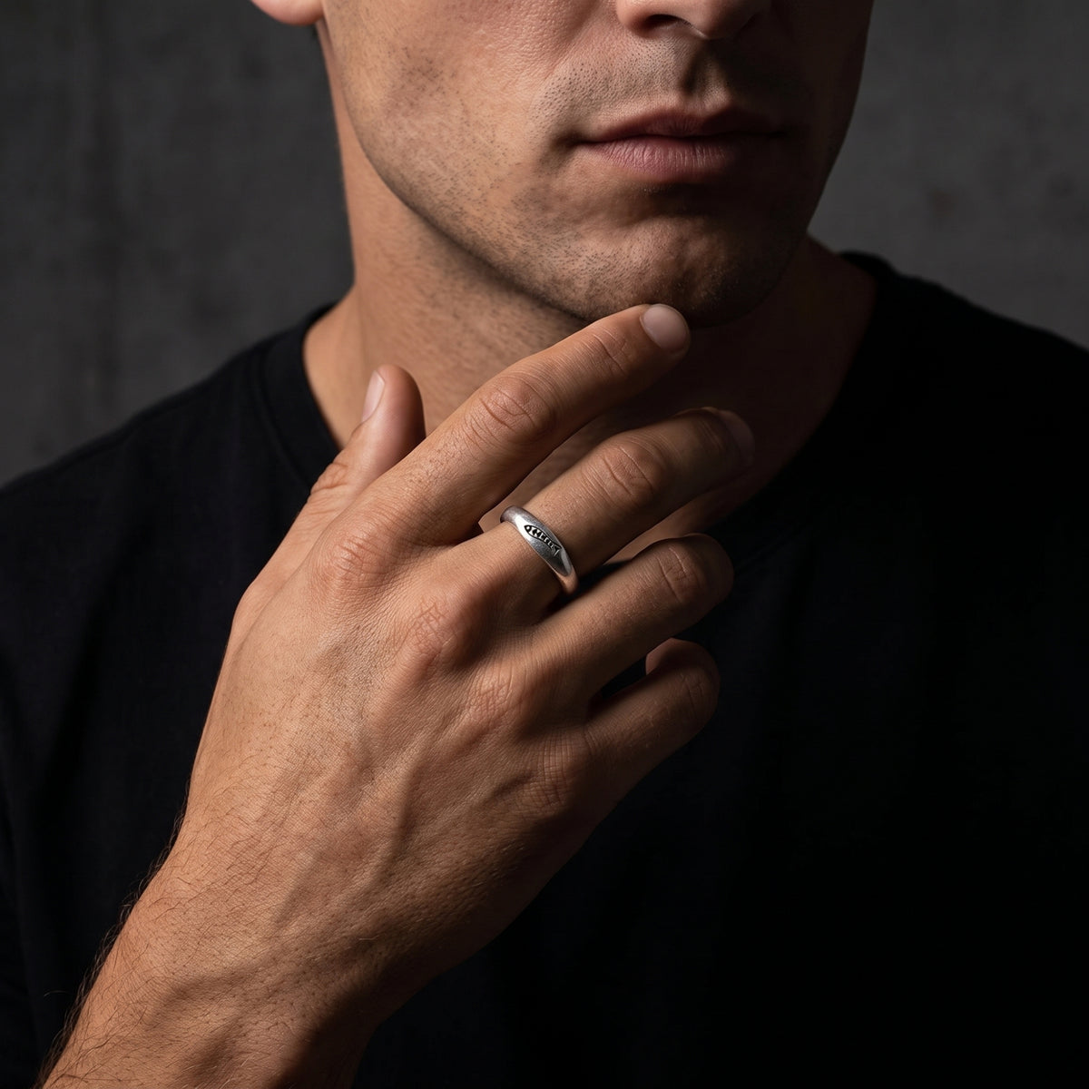Man wearing a silver carved fish ring with his hand resting against his chin over a black shirt.