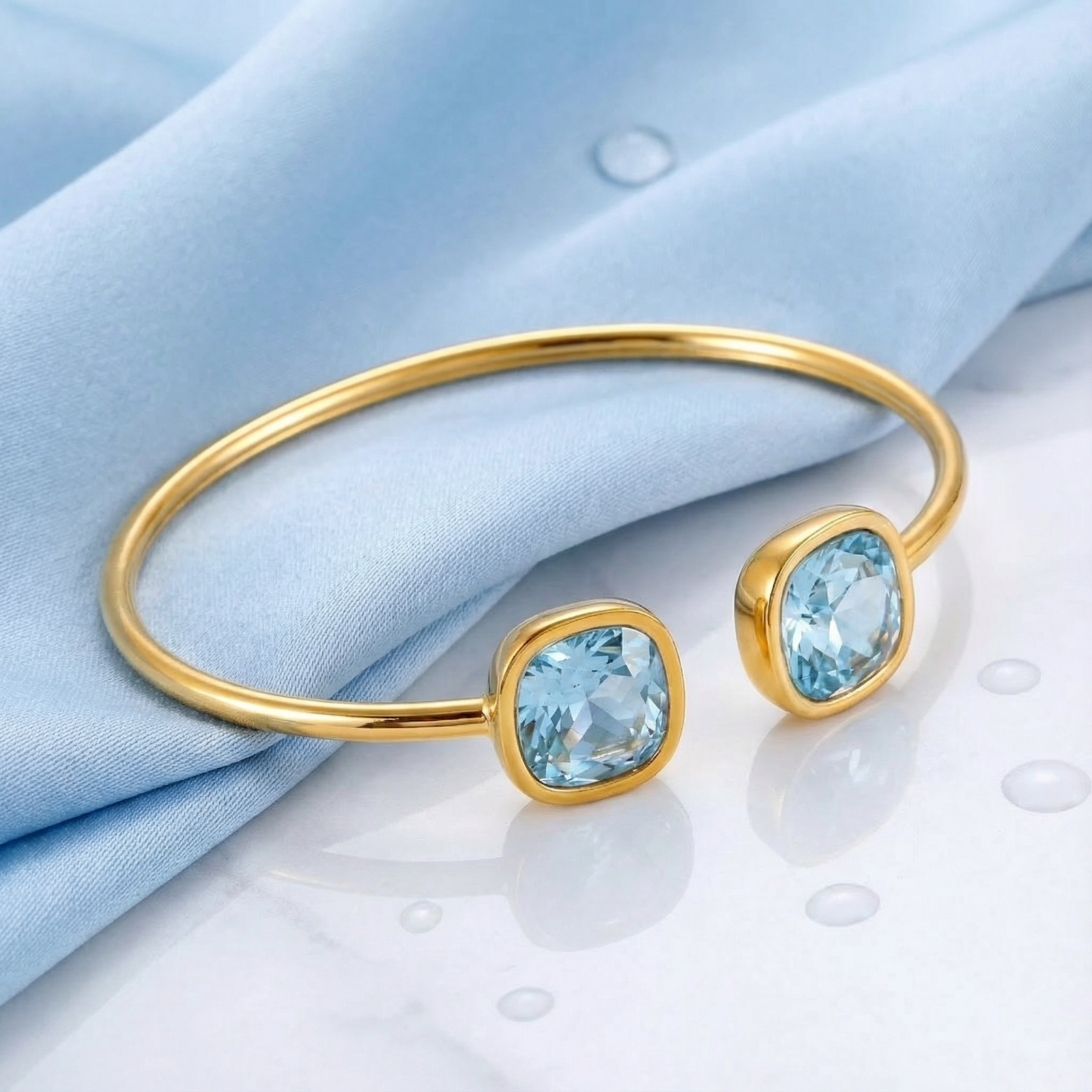 Gold bypass bangle with dual cushion-cut simulated aquamarine stones on light blue fabric and marble.