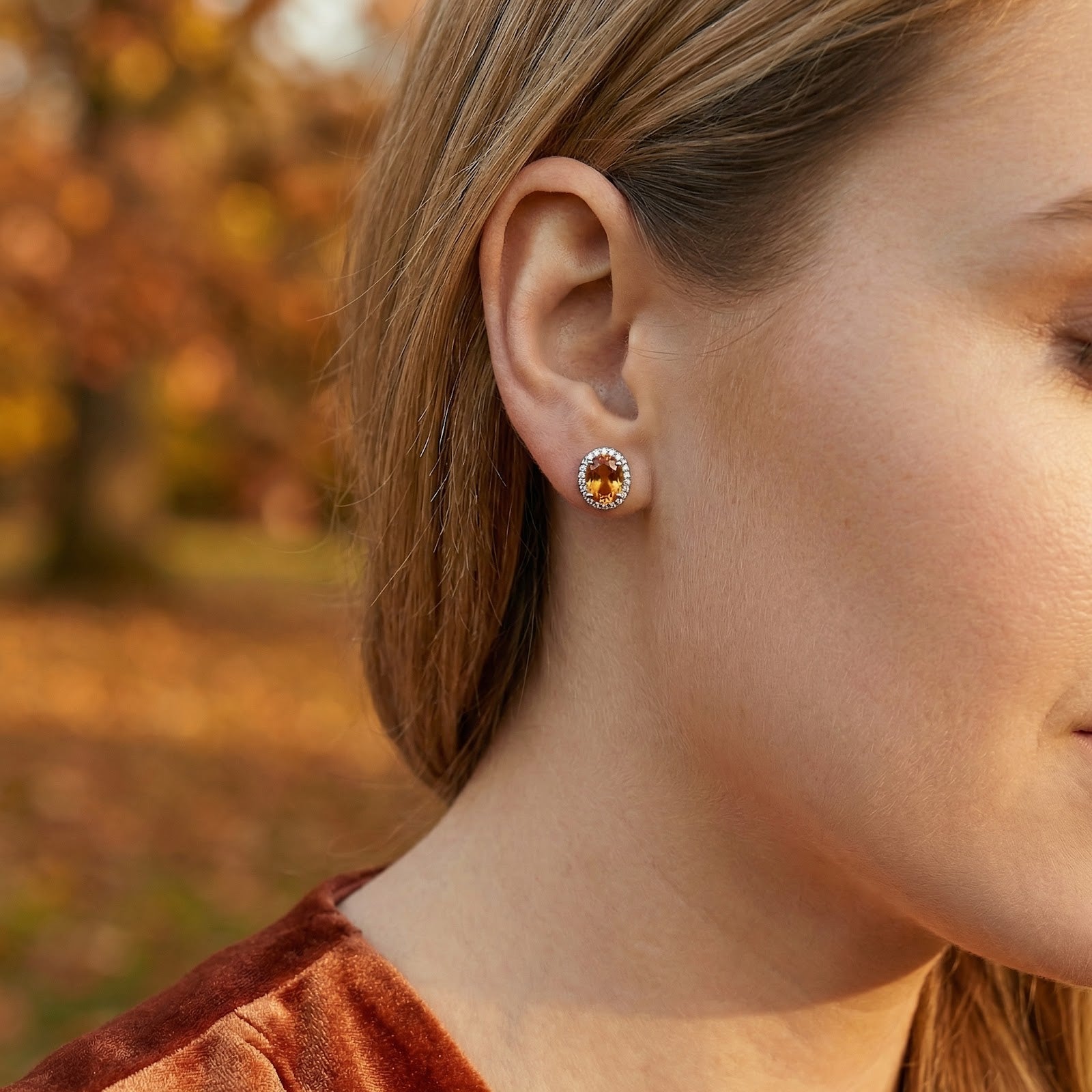 Woman wearing an oval simulated topaz halo stud earring in S925 sterling silver in an outdoor autumn lifestyle setting.