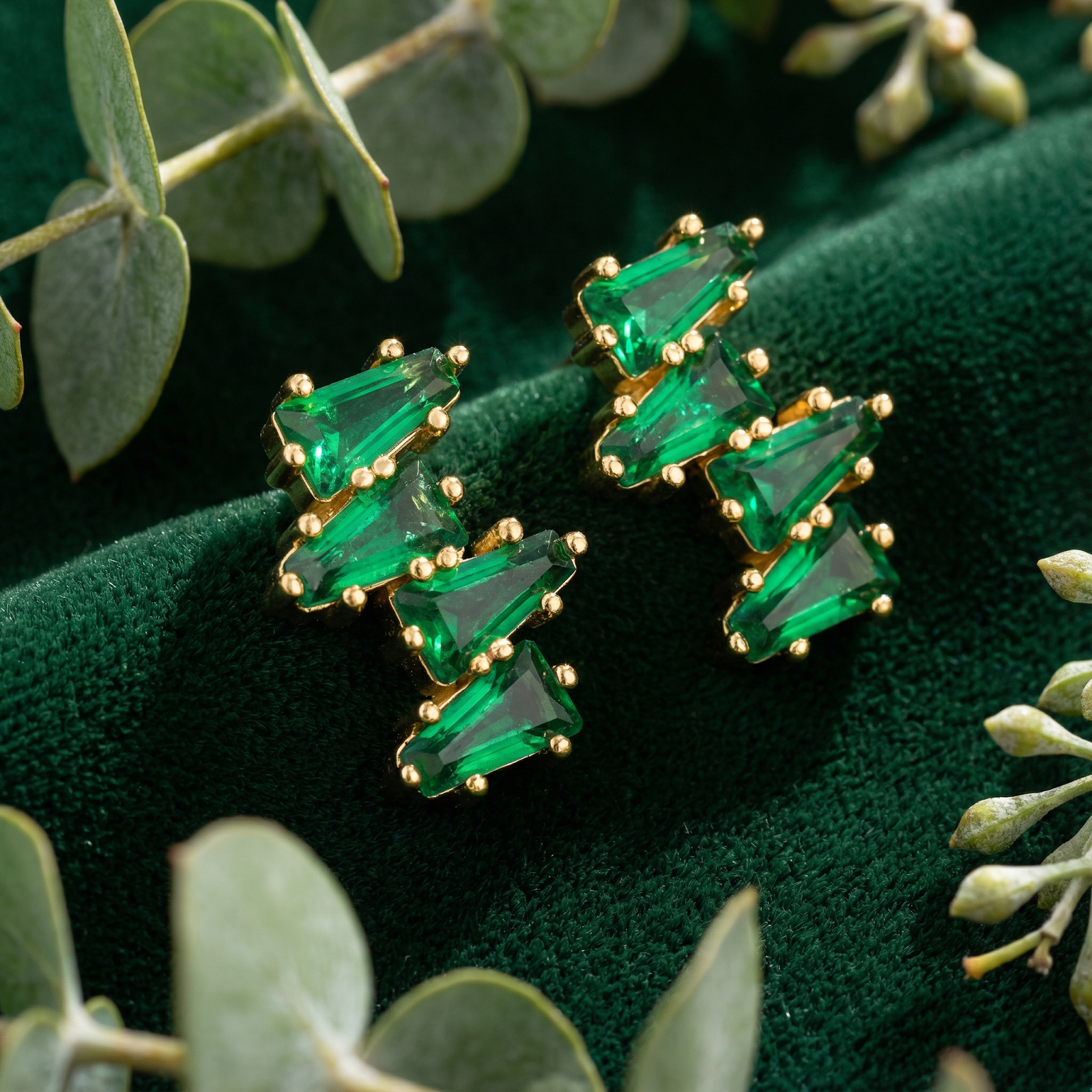 Macro view of geometric taper cut simulated emeralds in 18K gold vermeil earrings on green velvet.