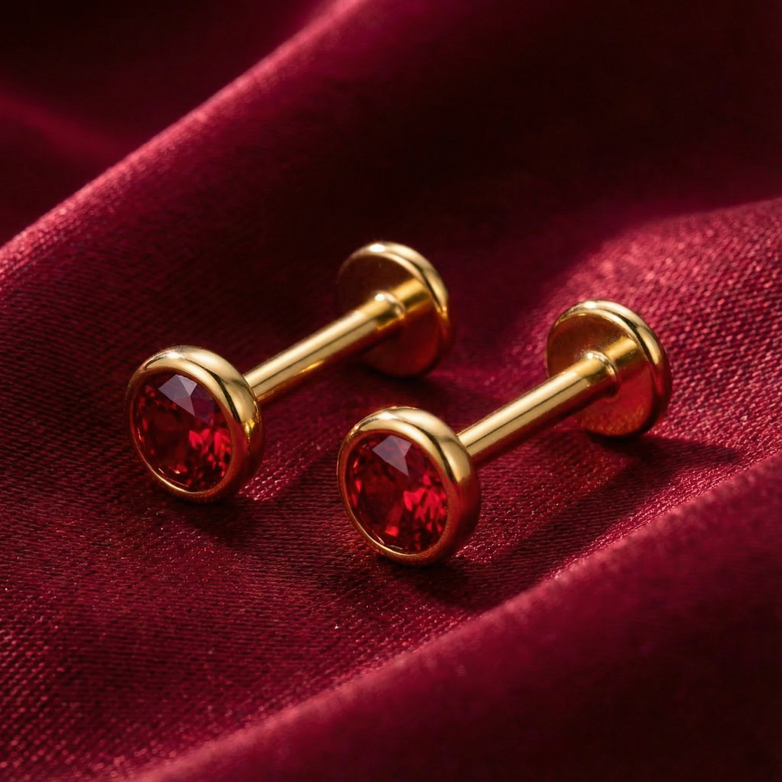 Side profile of gold vermeil labret-style stud earrings with round-cut simulated rubies on red fabric.