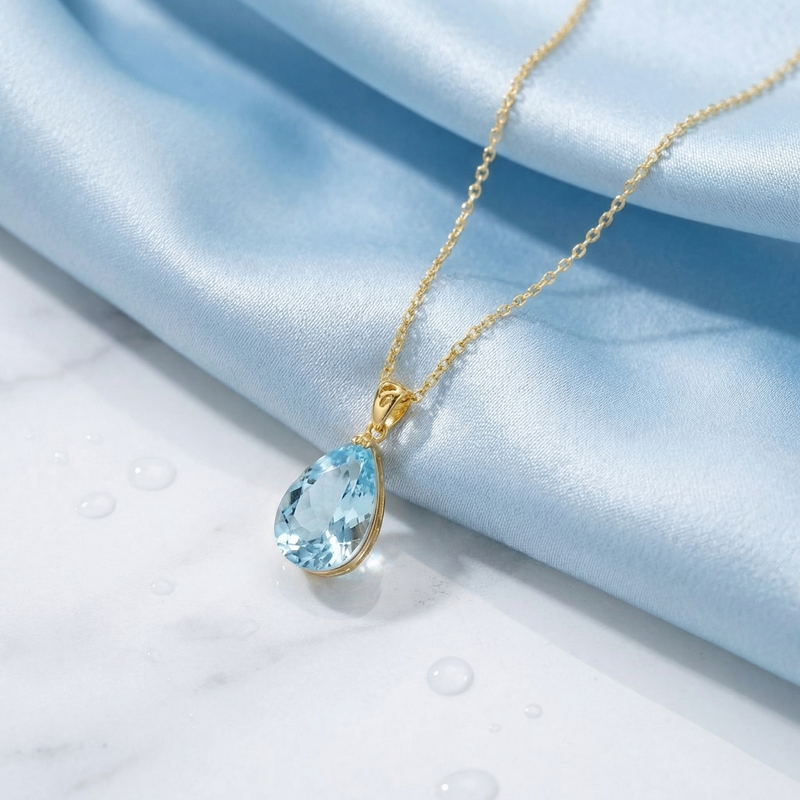 Side view of a pear cut simulated aquamarine necklace in 18K gold vermeil on blue silk fabric.