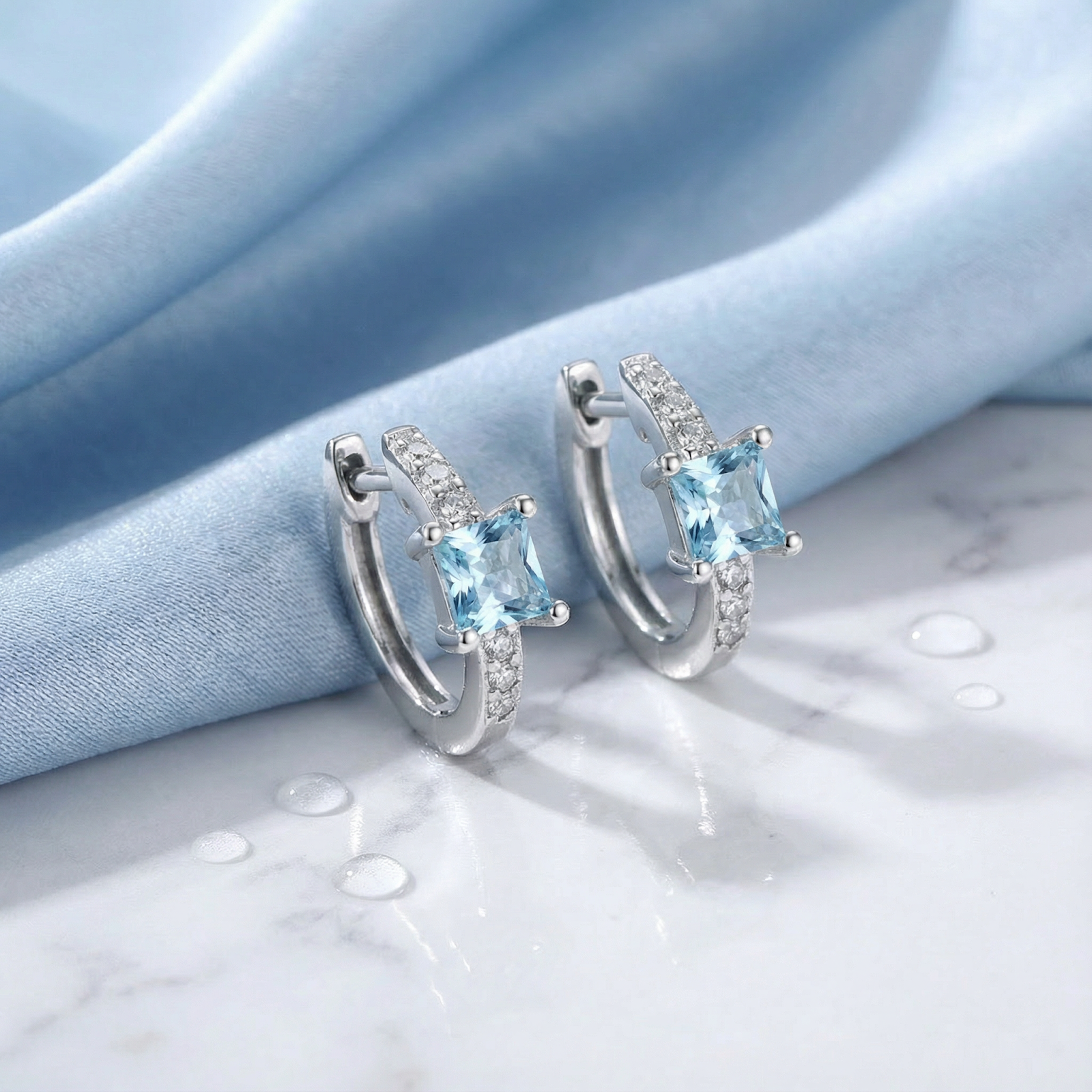 Sterling silver Aira huggie earrings with square light blue simulated aquamarine stones on blue fabric and marble.