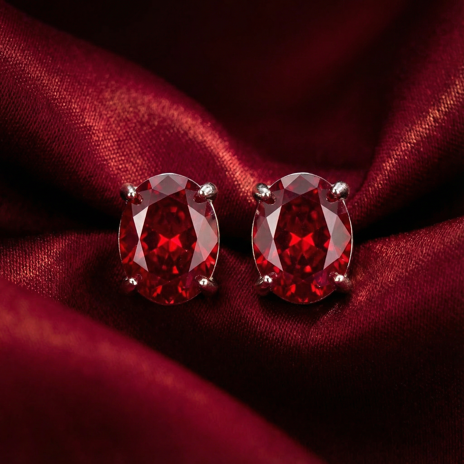 Close-up of oval-cut simulated ruby stud earrings in sterling silver settings against red fabric.