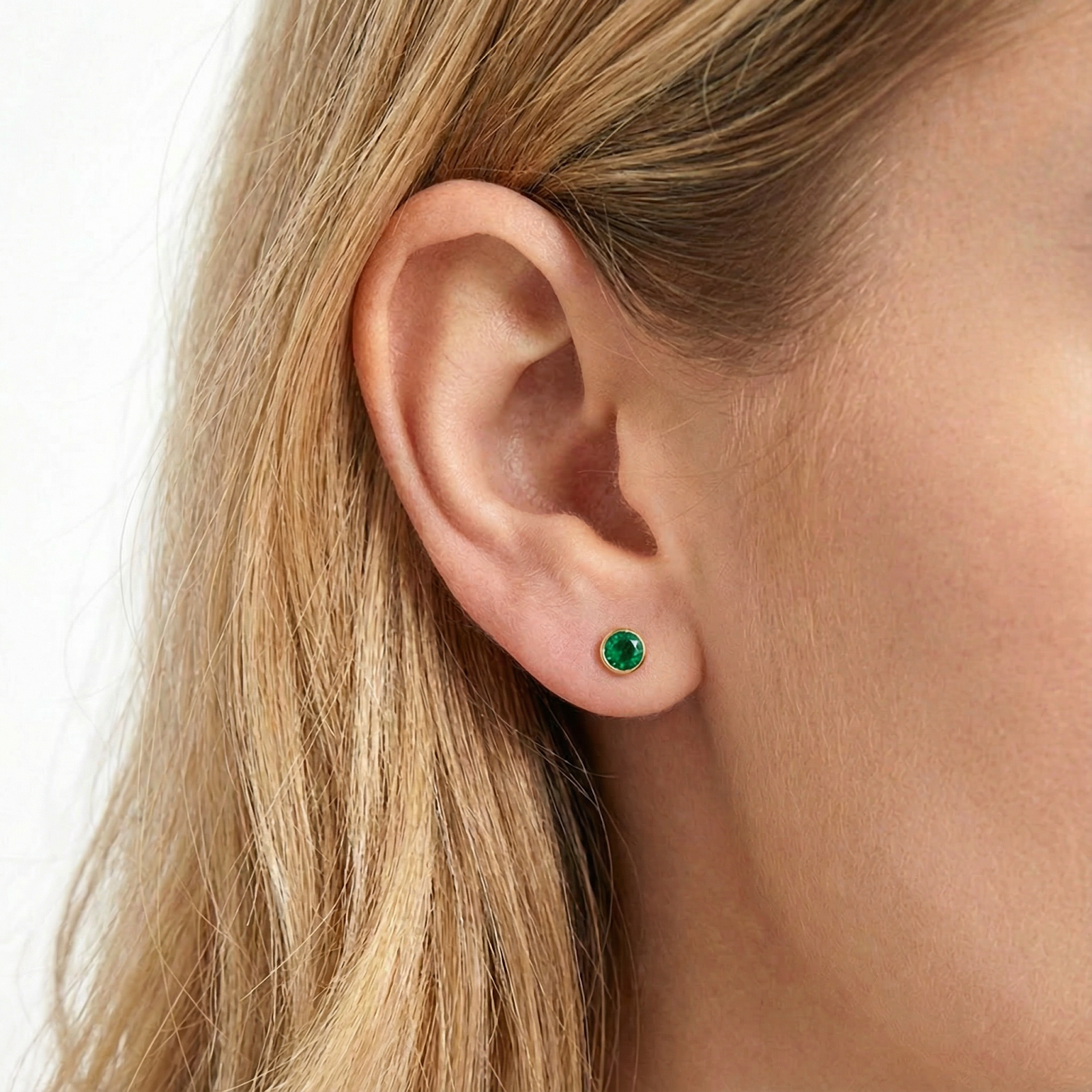 Macro view of a round simulated emerald bezel stud earring in 18K gold vermeil worn on a model's ear.