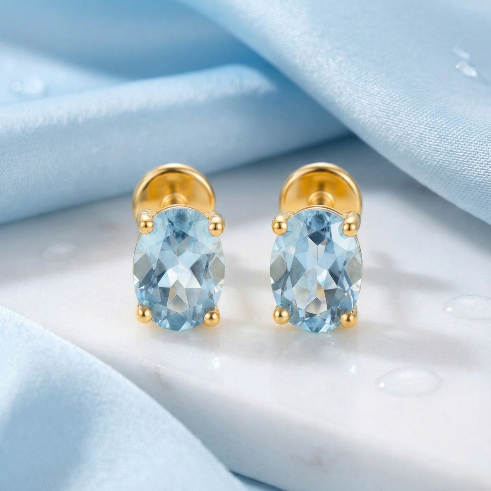 Front view of oval cut simulated aquamarine stud earrings in 18K gold vermeil on marble and blue silk.