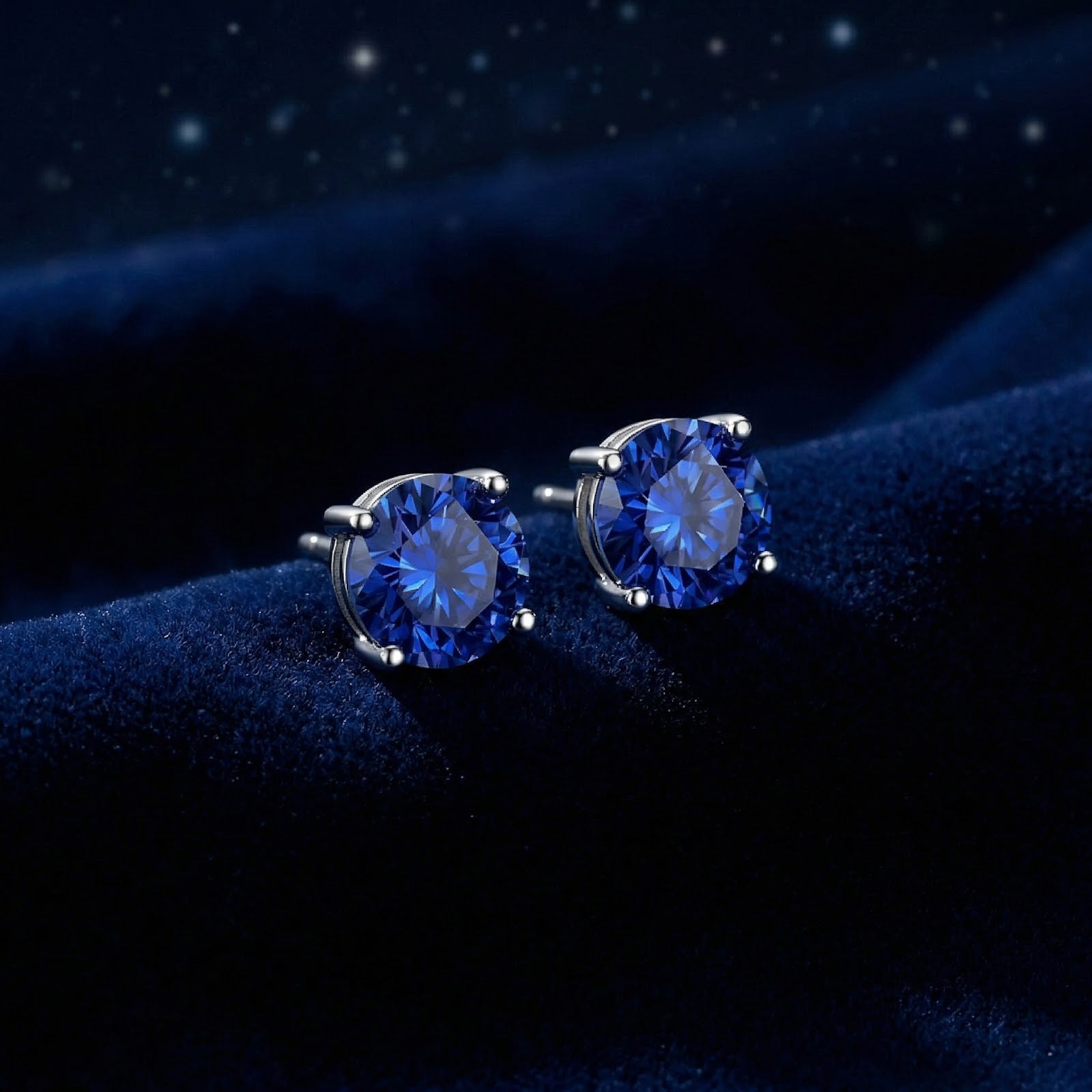 Side view of round blue sapphire stud earrings in silver settings resting on a textured blue velvet background.
