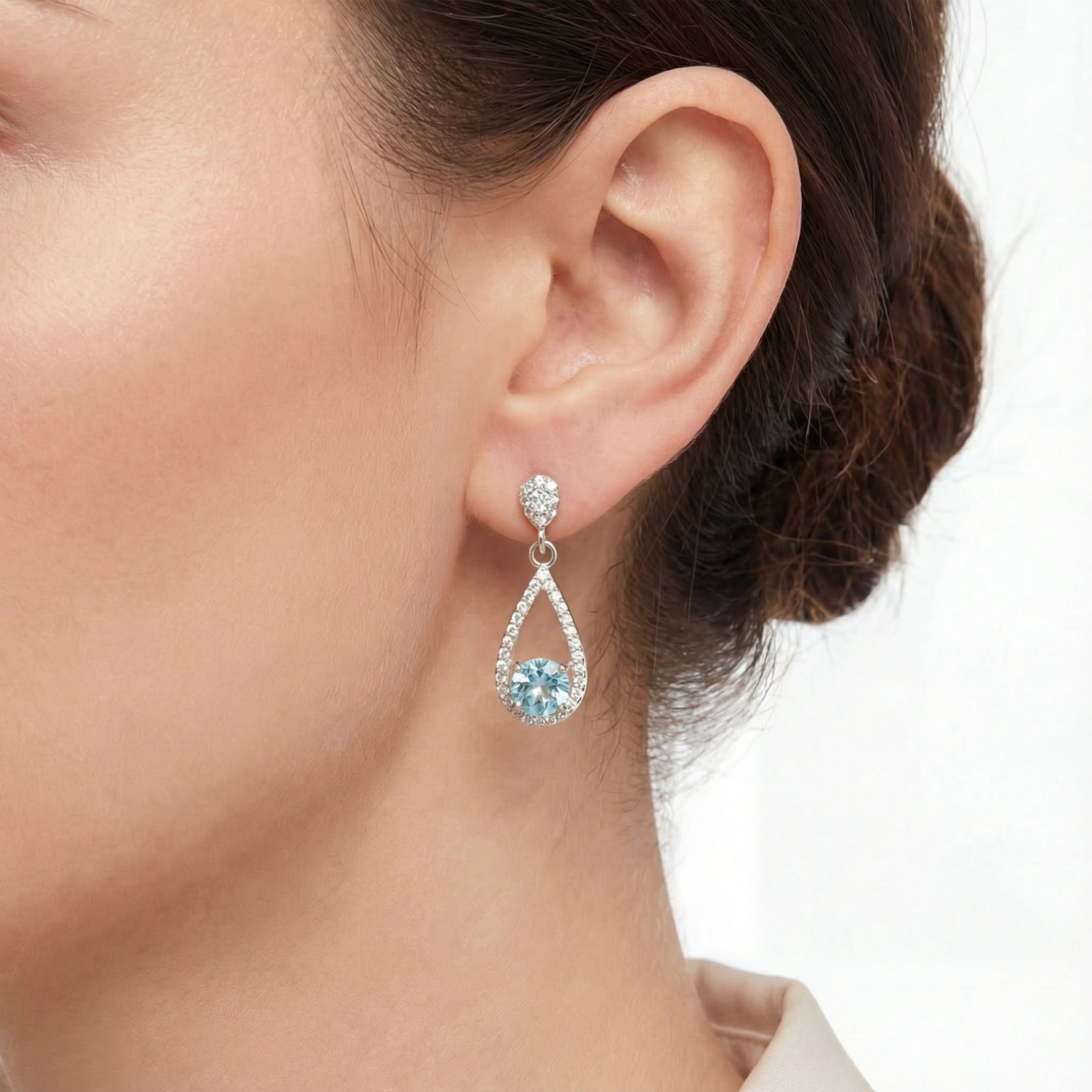 Close-up of a sterling silver teardrop drop earring with a round simulated aquamarine stone on a model's ear.