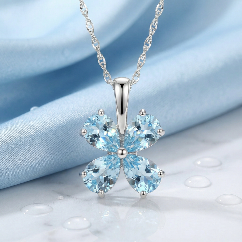 Macro view of a silver clover pendant with pear-cut simulated aquamarine stones on light blue silk with water droplets.