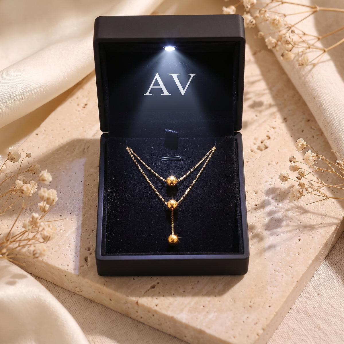 Gold layered lariat necklace inside a black illuminated gift box with the AV logo on a stone surface.