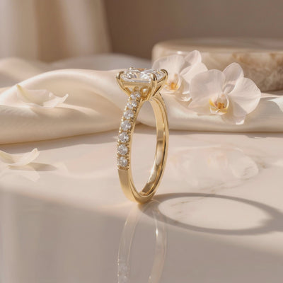 Side view of a radiant-cut solitaire ring with a pavé gold-tone band on satin beside white orchids.