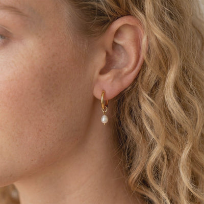 Close-up side view of a model wearing a gold-tone hoop earring with a dangling white pearl charm.