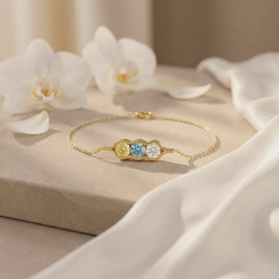 Gold-tone pea pod bracelet with yellow, blue, and clear gems laid flat on a stone surface near orchids.