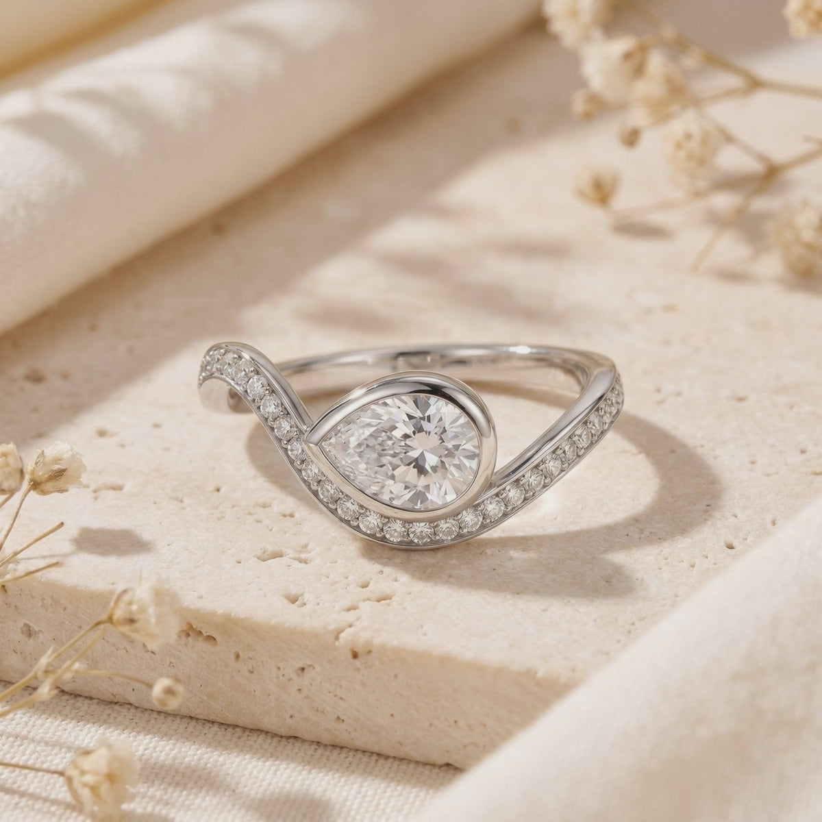 Close-up of a silver bypass ring with a bezel-set pear-cut moissanite resting on a beige stone surface.
