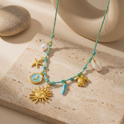 Turquoise woven charm necklace displayed on a beige stone slab with gold sun, moon, starfish, turtle, and shell charms.