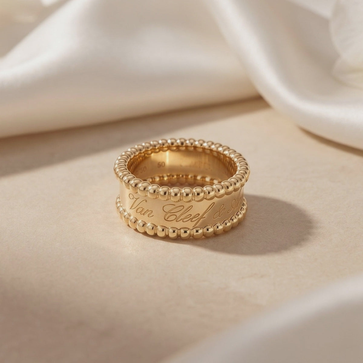 Gold-toned ring with script engraving and beaded borders standing on a beige surface beside white fabric.