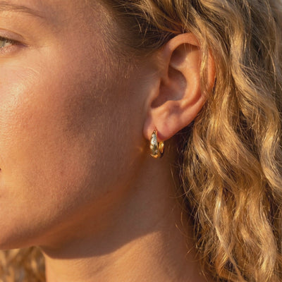 Side profile of a woman wearing a gold-tone starburst huggie hoop earring in warm sunlight.