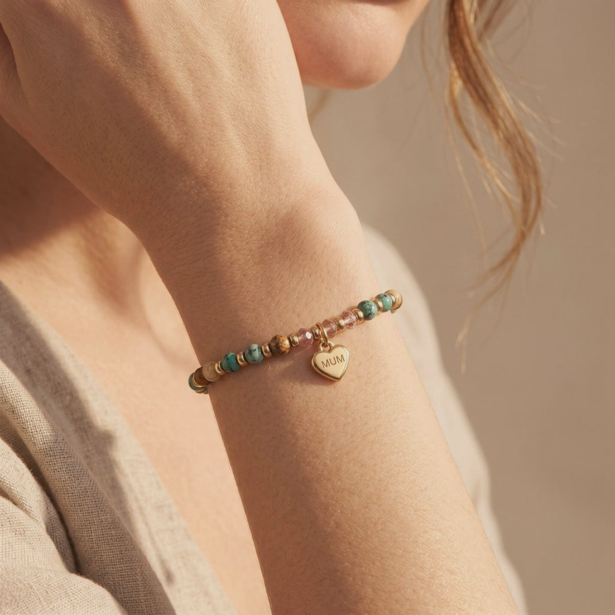 Gold-colored beaded bracelet with turquoise, jasper, pink crystal beads, and a MUM heart charm worn on a wrist.