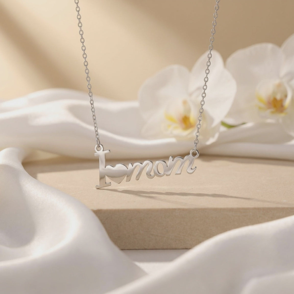Silver-finish “I [heart] mom” script necklace displayed over cream fabric beside white orchids.