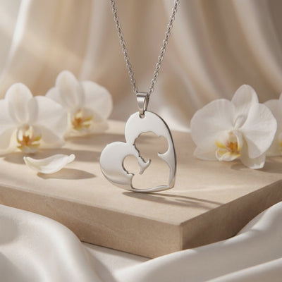 Silver heart cutout pendant with a mother and child silhouette, displayed upright on a pedestal among white orchids.