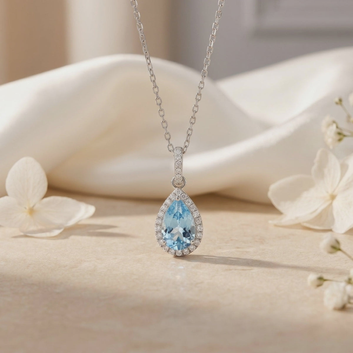 Silver necklace with a pear-cut light blue aquamarine halo pendant displayed near white flowers.