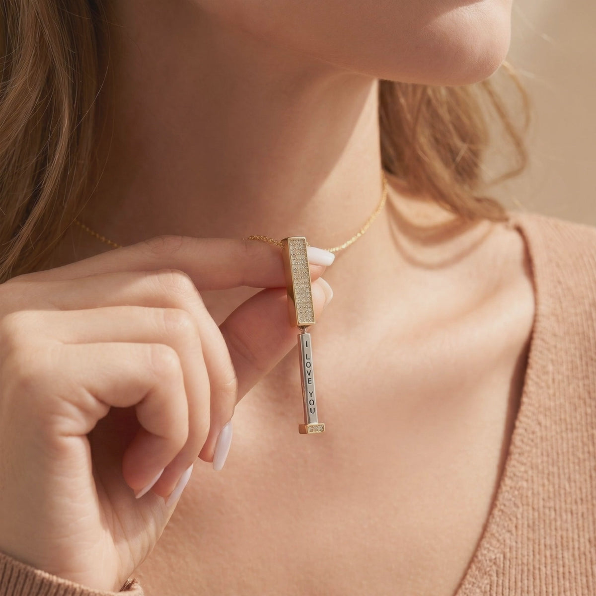 Woman holds a gold-tone necklace pendant open to show a vertical “I LOVE YOU” message beneath its crystal-paved bar.