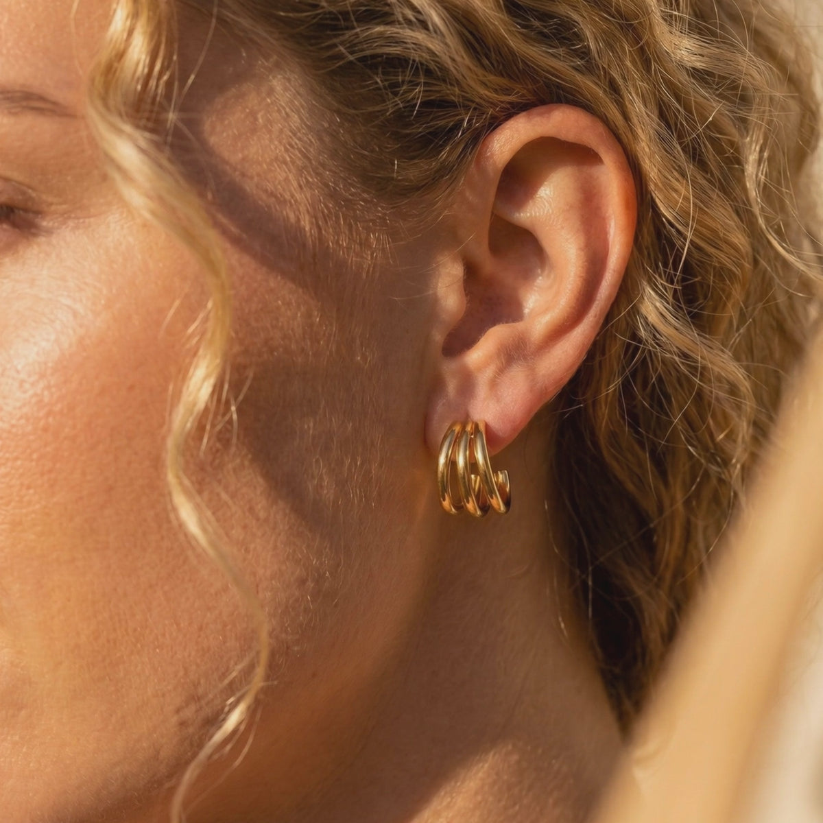 Gold-tone triple-band hoop earring worn on a model's ear, shown against curly blonde hair.