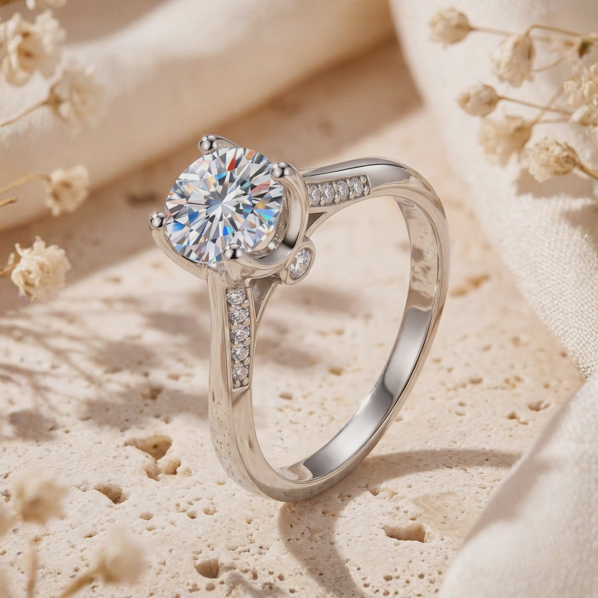 Close-up side view of a silver solitaire ring with a round center stone and underside accent stone on a porous surface.