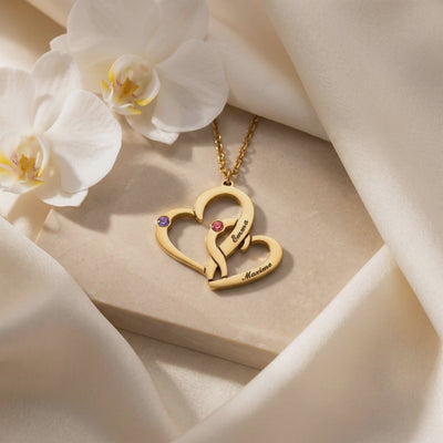 Close-up of the gold-tone double-heart pendant engraved with Emma and Maxime, with purple and pink stones, beside orchids.