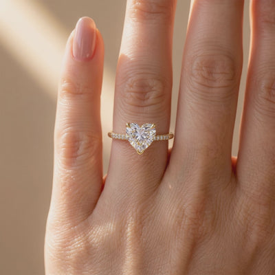 Close front view of a heart-cut solitaire ring with a pavé band in yellow-toned metal, worn on a finger.
