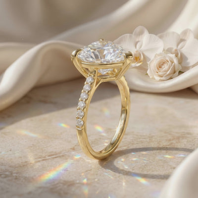 Side view of a gold-tone heart-cut clear-stone ring with a pavé band standing upright beside white flowers