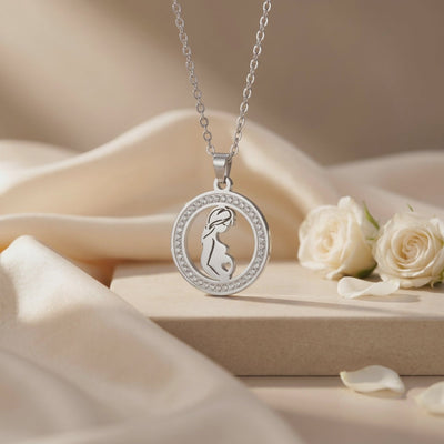 Silver pregnancy silhouette pendant with a crystal halo, suspended over draped fabric beside white roses.