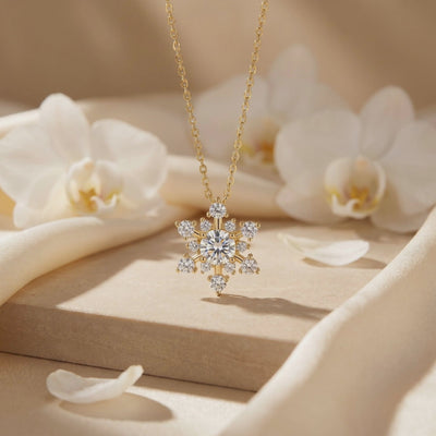 Gold-tone snowflake pendant necklace hanging above a beige pedestal, with white orchid flowers in the background.