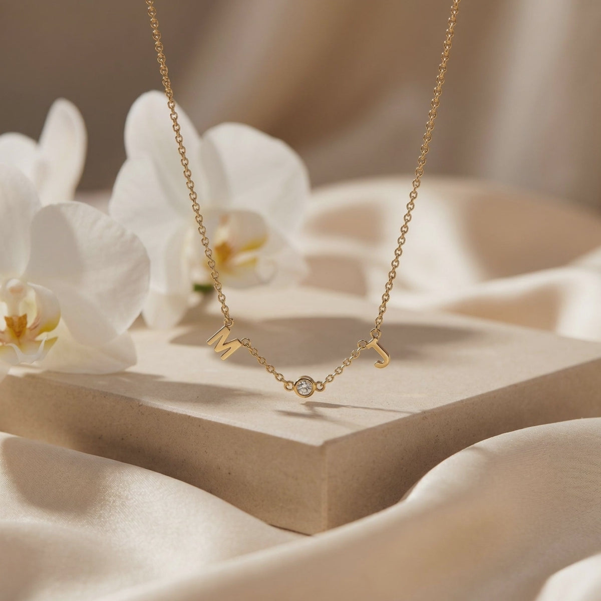 Gold-tone M and J initial necklace with a bezel-set round accent draped over a neutral display block.