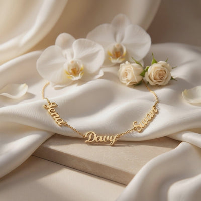 Gold-tone necklace with three script name charms displayed on draped white fabric beside white orchids and roses.