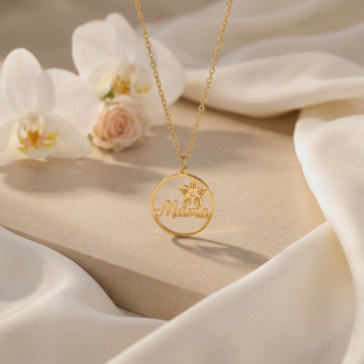 Gold-tone circular Mama pendant necklace displayed on a beige block with white orchids