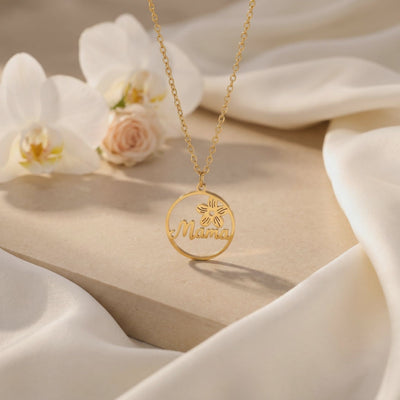 Gold-tone circular Mama pendant necklace displayed on a beige block with white orchids