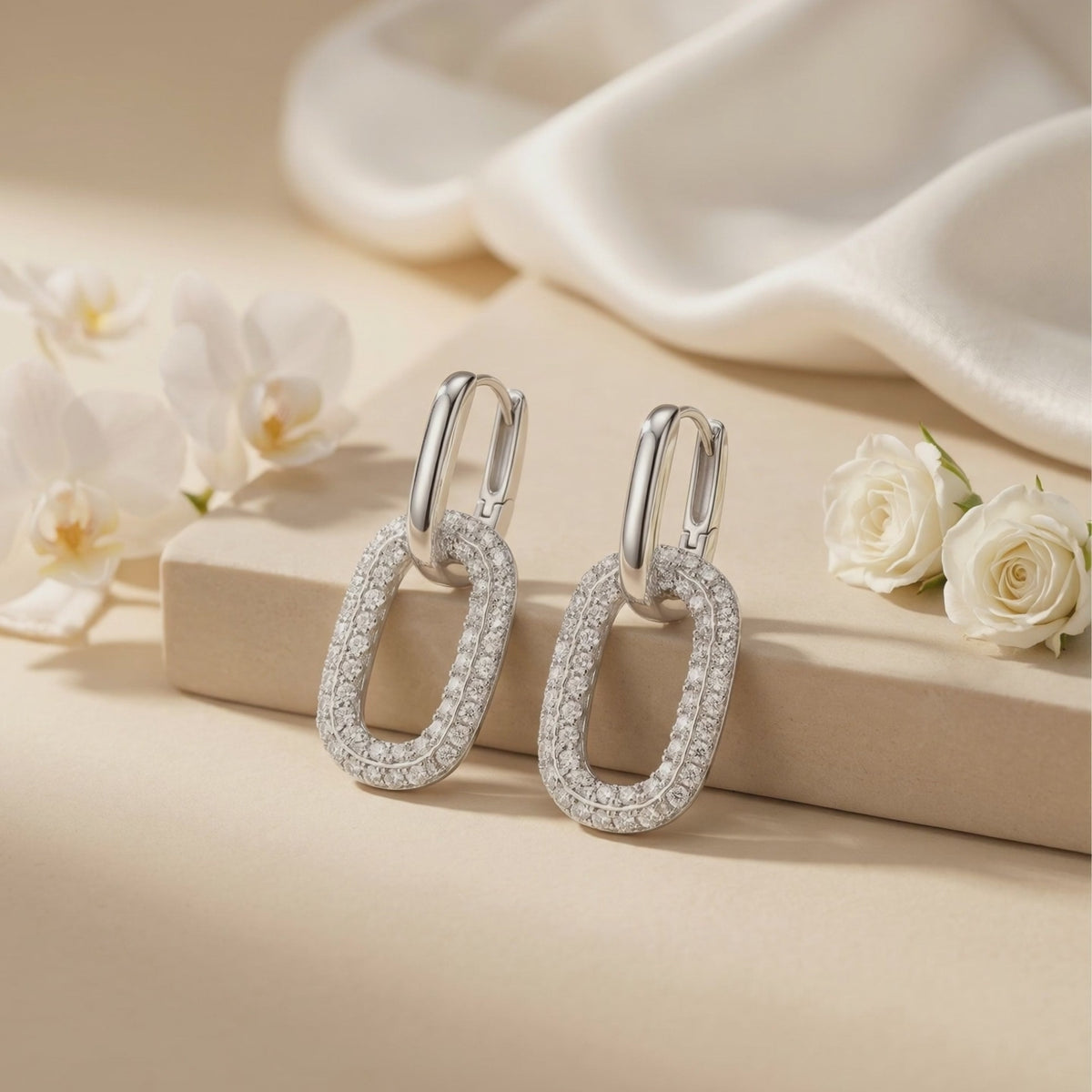 Pair of silver double-link moissanite earrings displayed on a beige block near white roses.