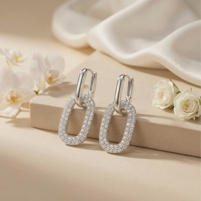 Pair of silver double-link moissanite earrings displayed on a beige block near white roses.