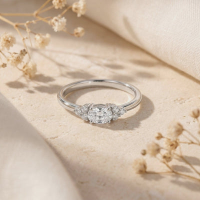 Silver oval moissanite ring with clustered side accents, displayed on a textured beige surface with dried flowers.