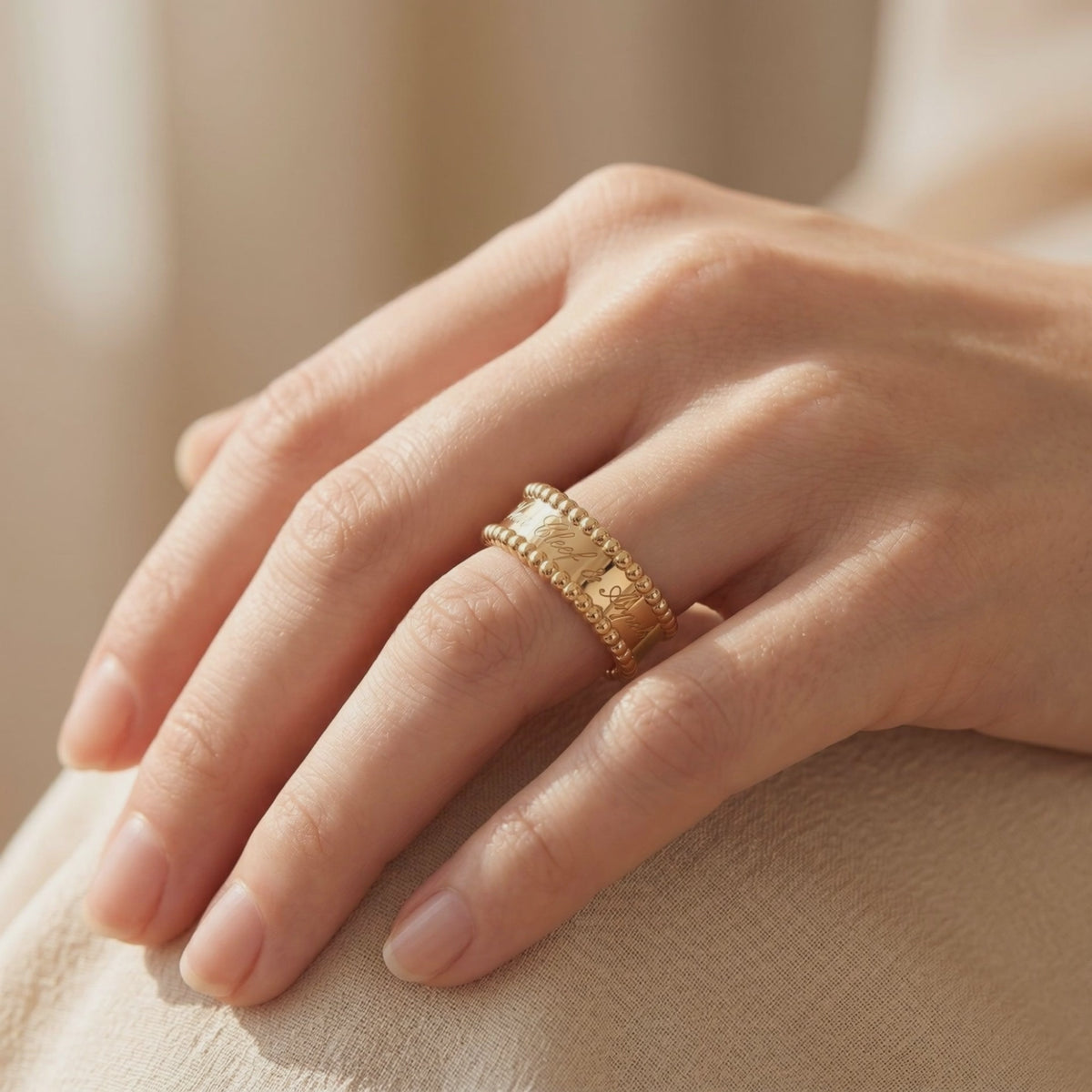 Wide gold-toned ring with script engraving and beaded borders worn on a finger.