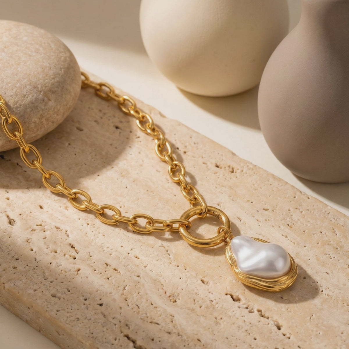 Gold-toned link necklace with an irregular white pearl pendant resting on a textured beige slab.