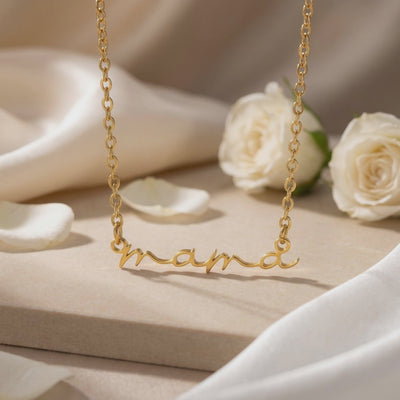 Gold-toned script mama necklace displayed on a beige pedestal with white roses.