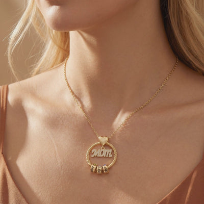 Woman wearing a gold-tone MOM necklace with a twisted circle frame, heart detail, and engraved name rollers.