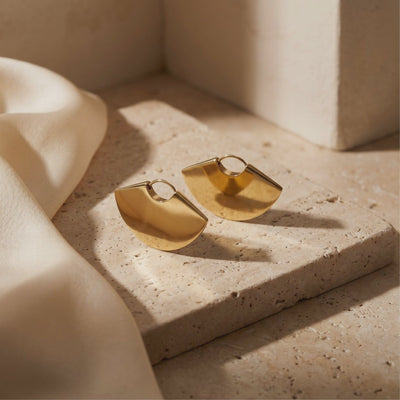 Pair of gold-tone fan-shaped earrings displayed on a textured beige stone block beside draped cream fabric.