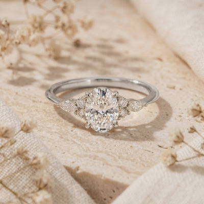Silver oval moissanite ring with cluster side stones resting on a beige stone surface.