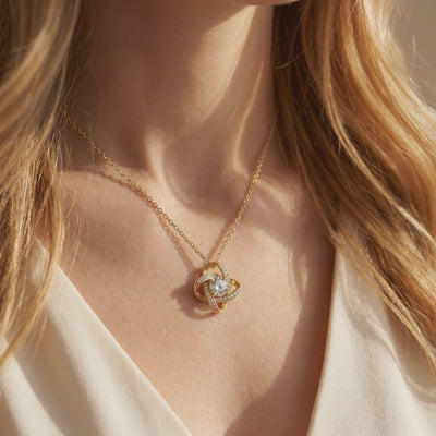 Gold-toned interlocking knot necklace with a clear center stone, shown in a close-up while worn on a person's neck.